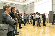 Rund 20 Personen stehen und hören einem Vortragenden zu, der in diesem Bild allerdings nicht zu sehen ist.