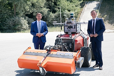 Markus Achleitner und Jochen Danninger stehen links und rechts neben einem automatisiertem Nutzfahrzeug. Foto: Land OÖ/Liedl